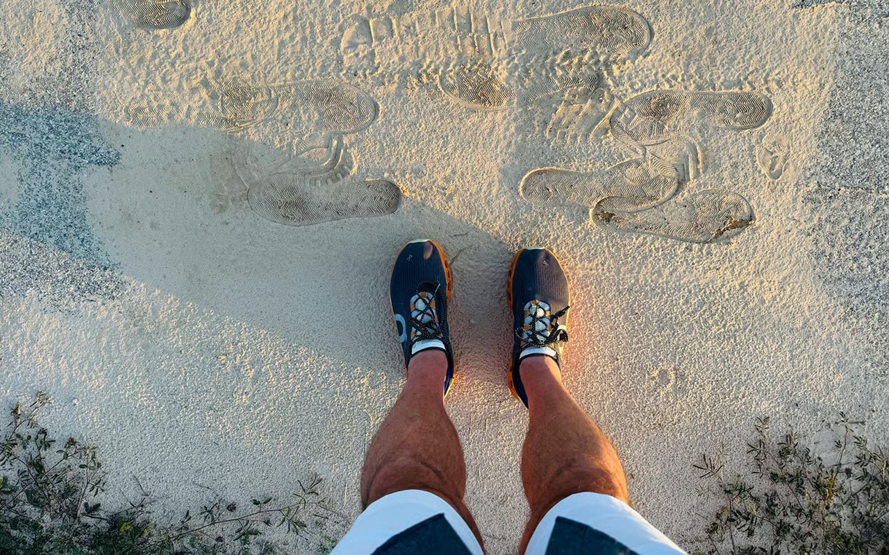 A person stands on pale sand in athletic shoes among overlapping footprints.