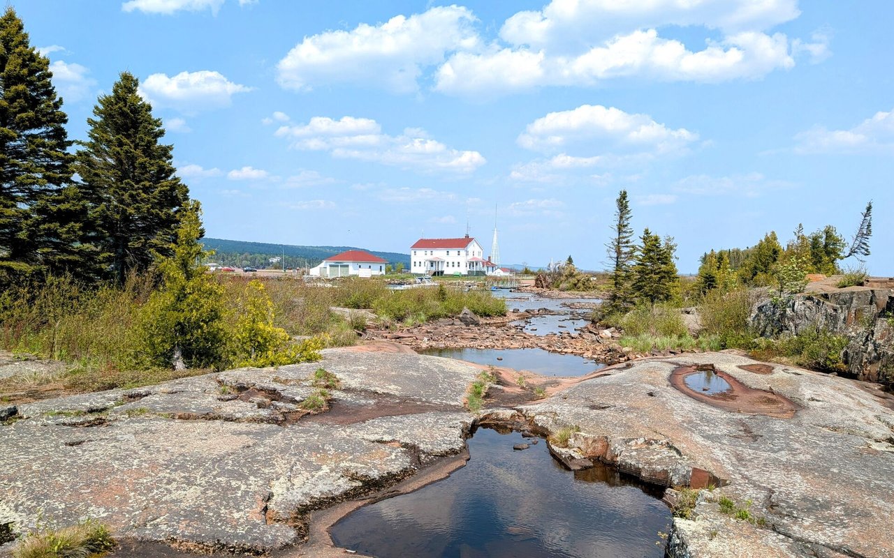Rocky North Shore shoreline with a shallow stream, evergreen trees, and distant buildings.