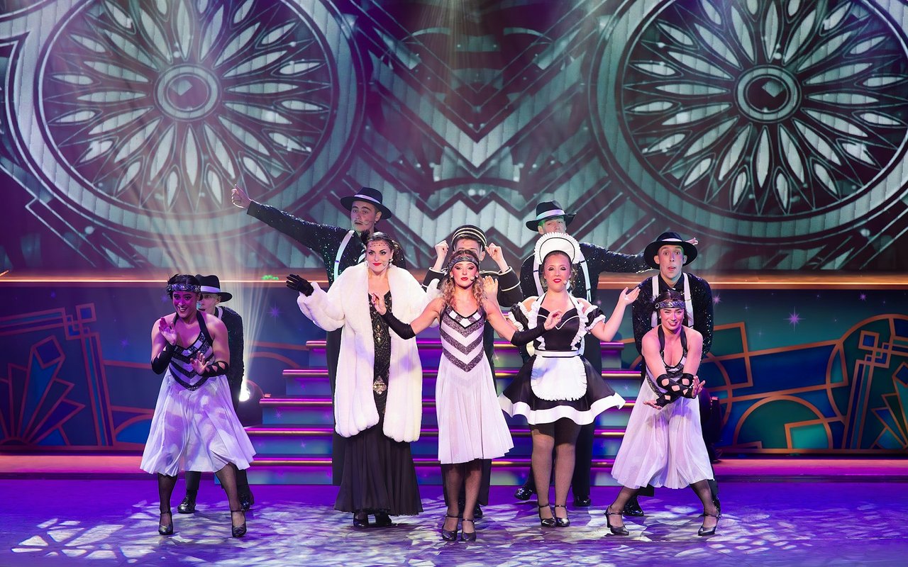 Nine Roaring Twenties-costumed performers pose on a stage with an Art Deco backdrop.