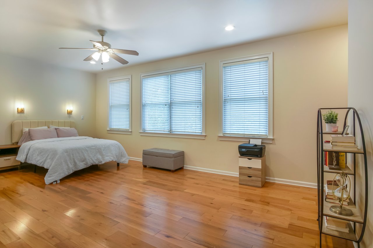 Spacious bedroom with a large bed and light blue bedding. Wooden floor, three windows with blinds, and a ceiling fan. Calm and cozy atmosphere.
