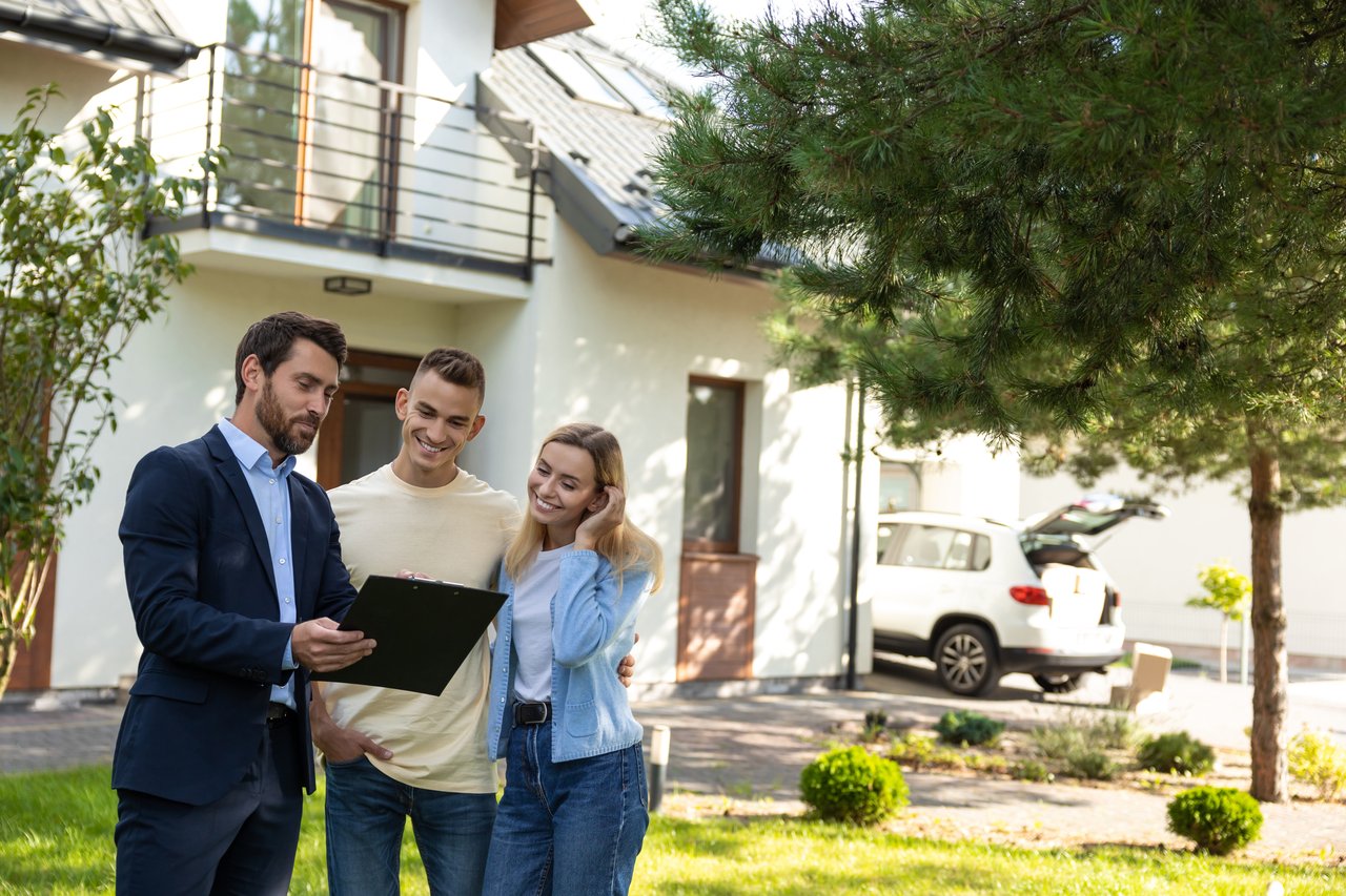 Real estate agent shares a clipboard with a smiling couple outside a modern home.