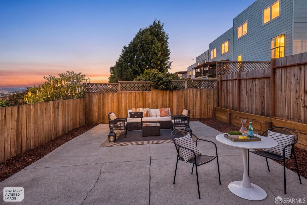 Furnished concrete patio with outdoor dining and lounge seating beside a tall wood fence at sunset.