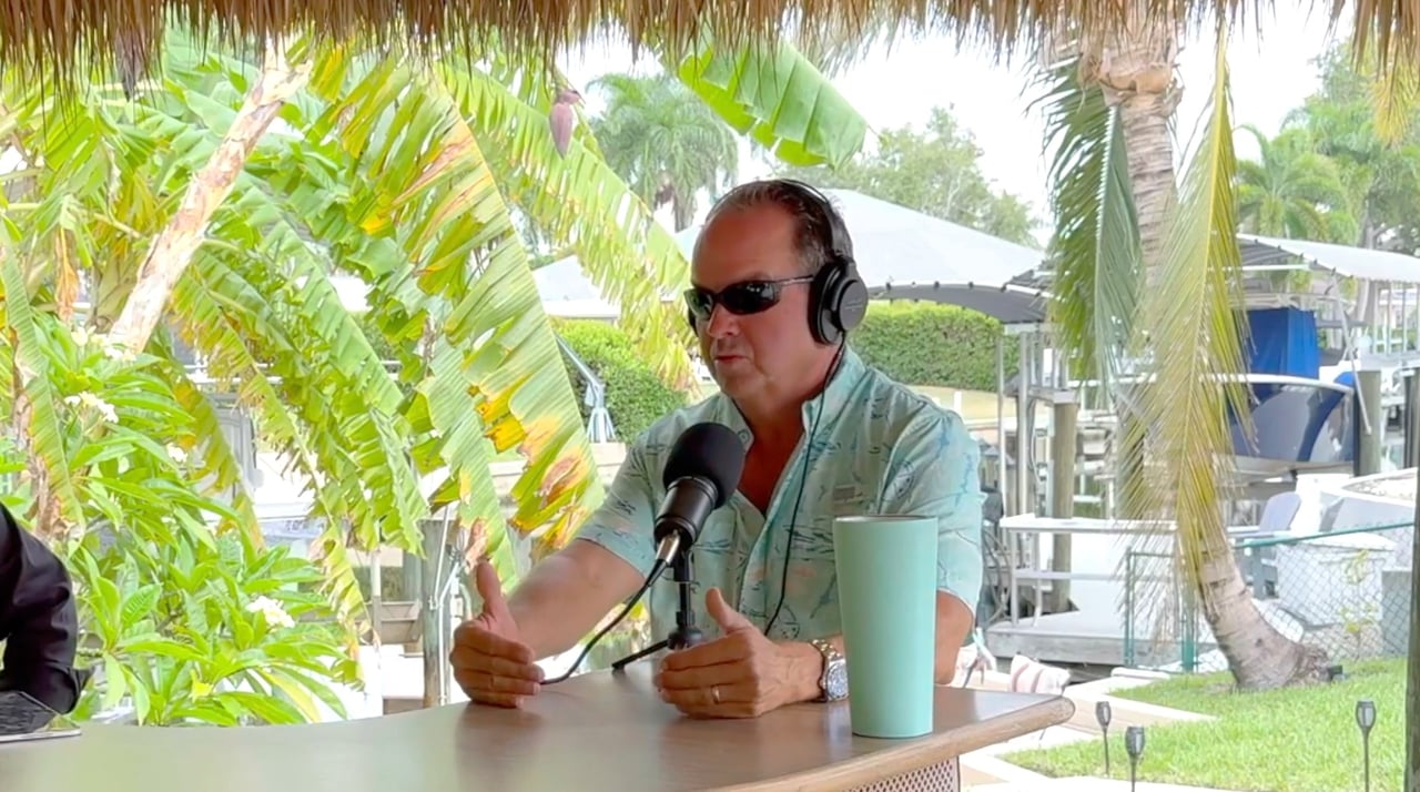 Man wearing headphones speaks into a microphone at a tropical waterfront table.