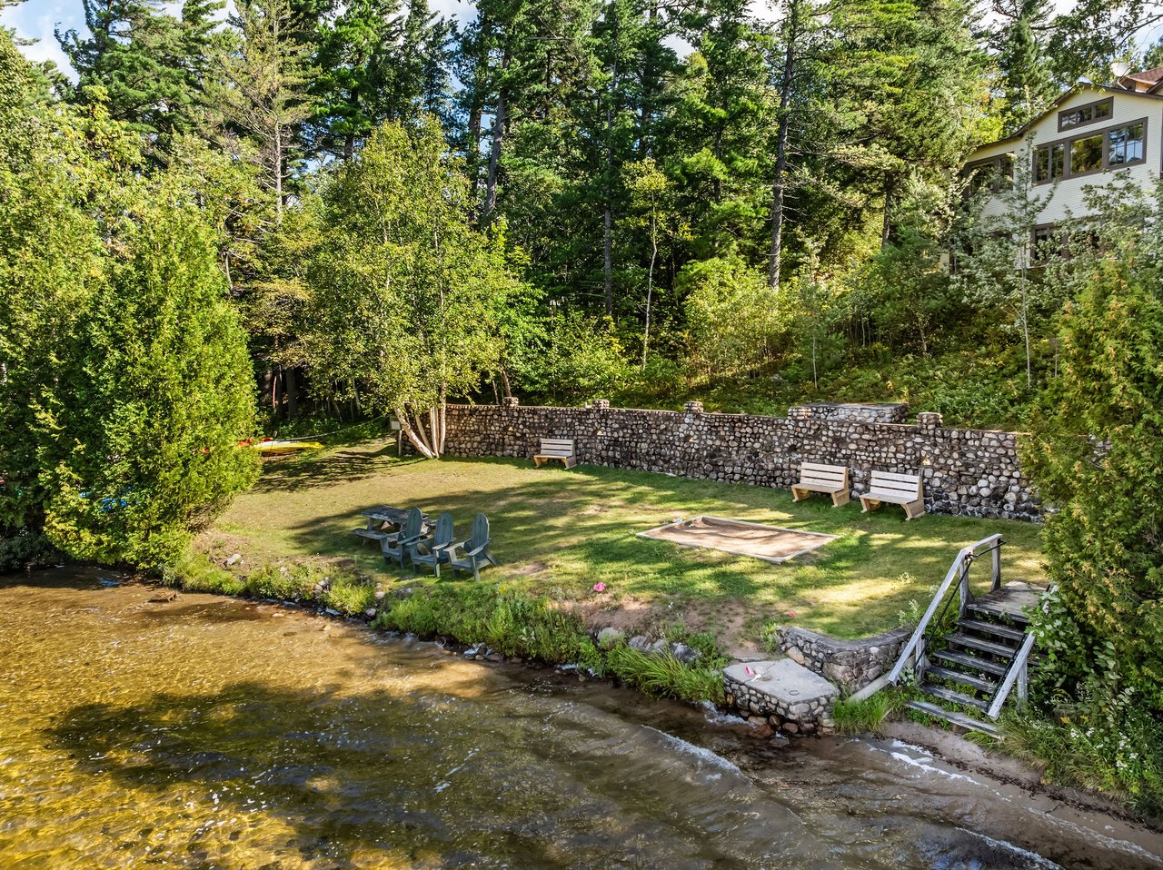 Riverside yard with a stone wall, fire pit, outdoor seating, and wooded house
