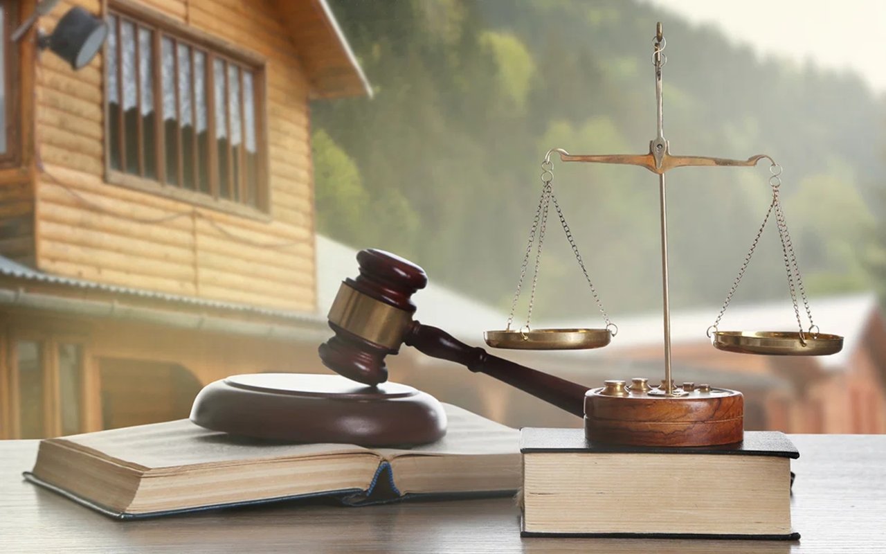 Scales of justice and a gavel on law books, with a wooden house behind them.