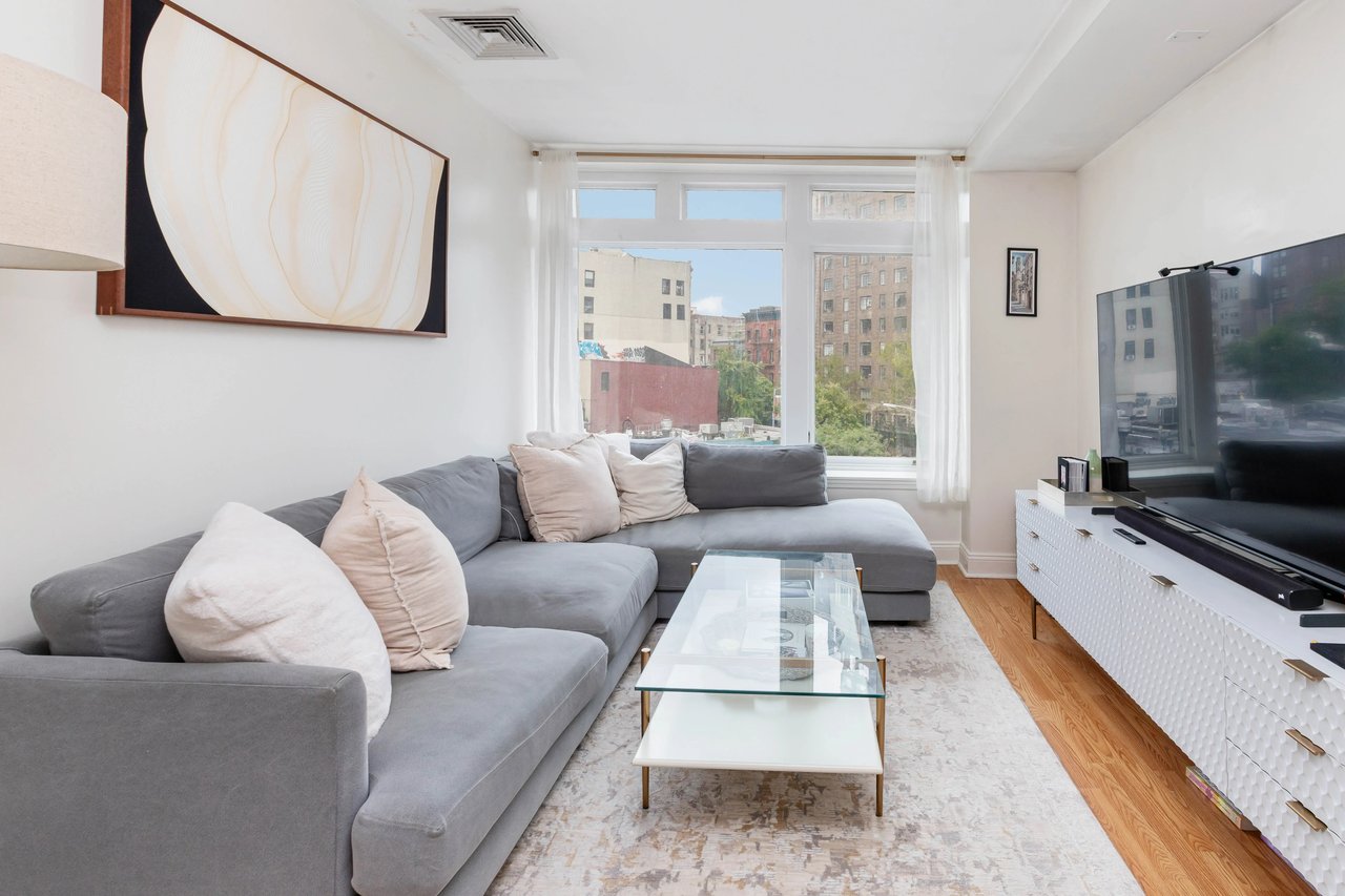 Furnished living room with a gray sectional, glass coffee table, and large windows overlooking city buildings.