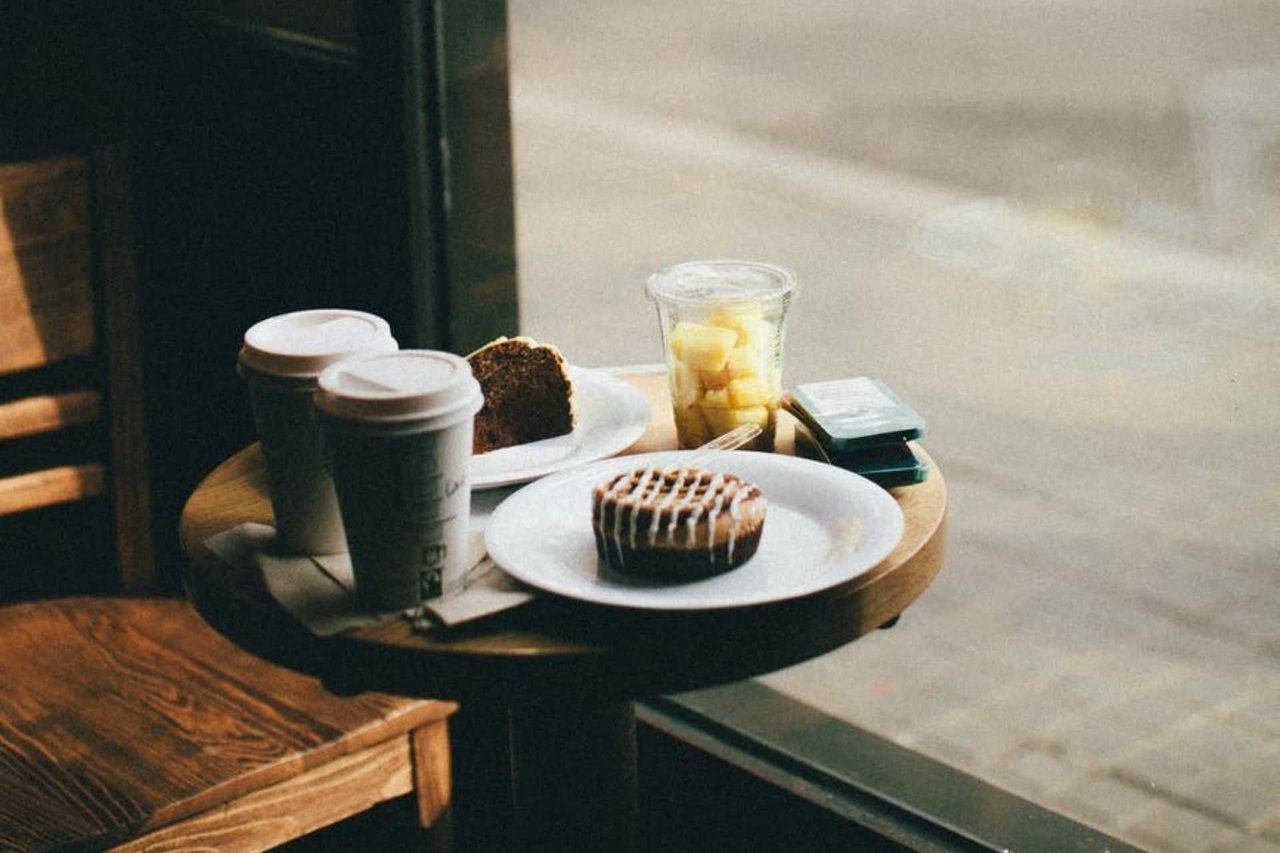 Cozy morning cafe setup featuring breakfast pastries, a drizzled cinnamon roll, a slice of banana bread, diced fresh fruit, and takeaway coffee on a wooden table.
