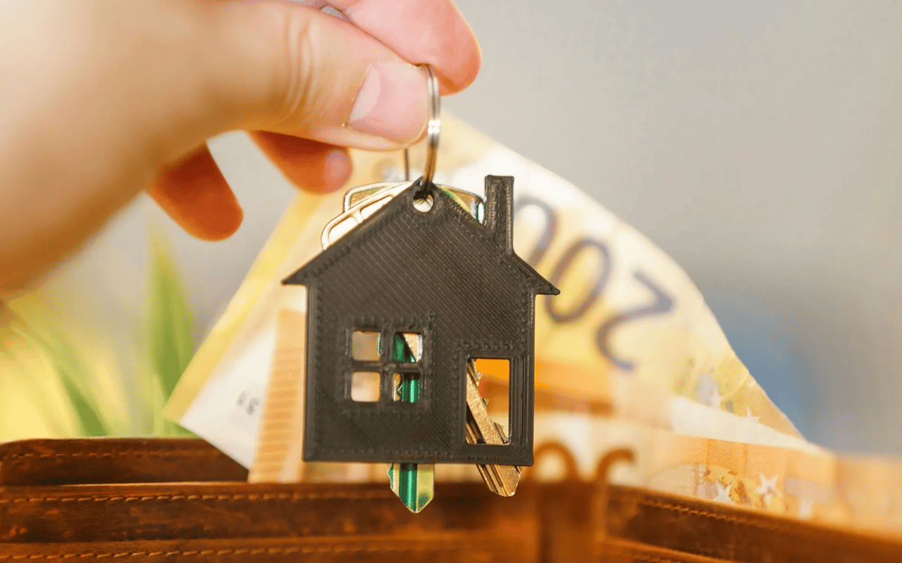 A hand holds house-shaped keys above banknotes in a brown leather wallet.