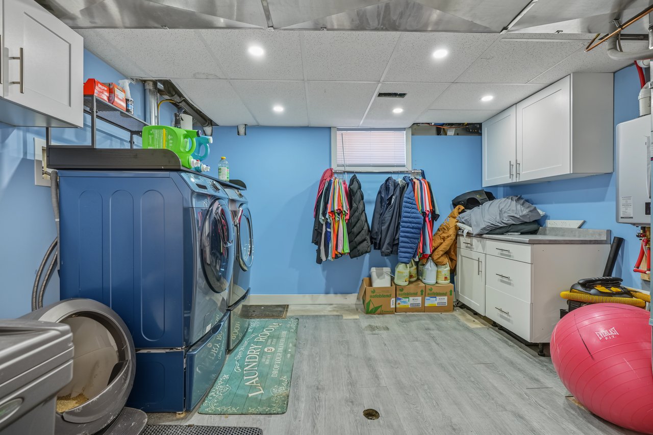 Laundry room with multiple machines, white cabinets, and clothes hanging beneath a window.