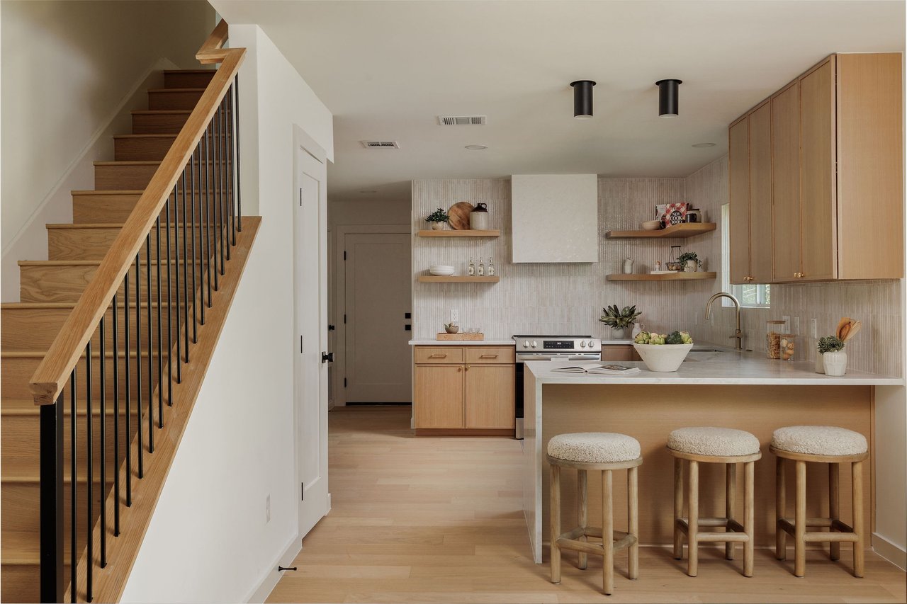 Kitchen with a white island and three stools beside a light wood staircase.