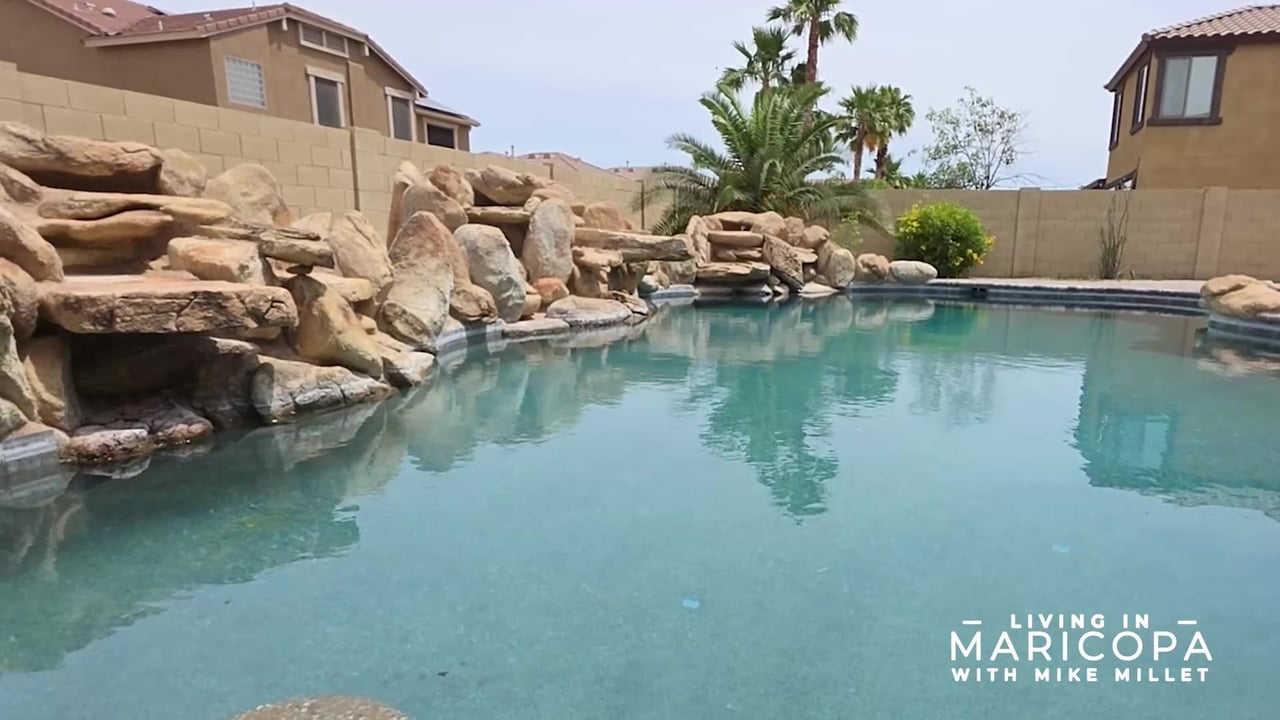 Backyard pool with a large rock waterfall, palm trees, and a tan block wall.