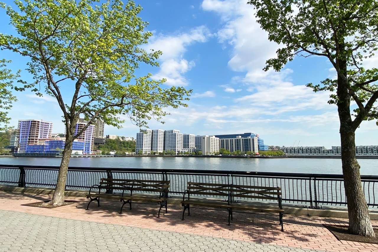Hoboken waterfront promenade with benches, leafy trees, and buildings across the river.