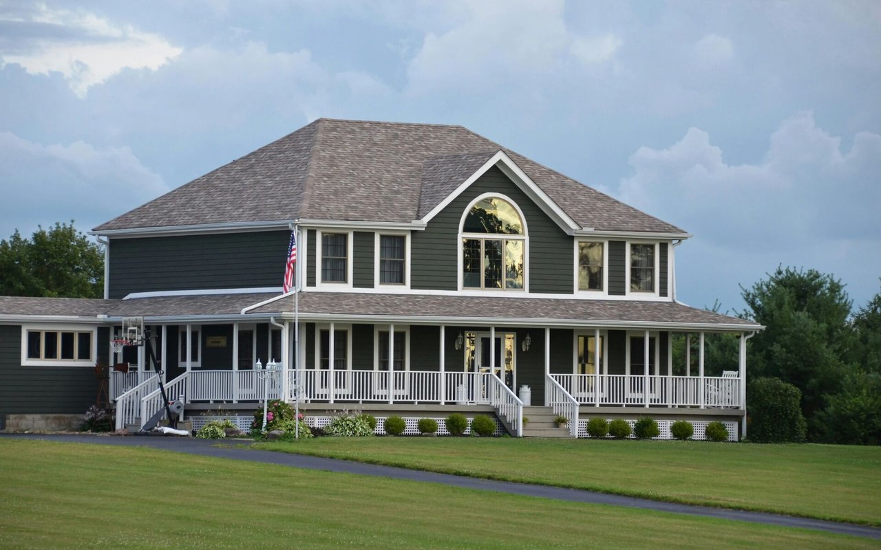 Dark green two-story house with a white wraparound porch and arched upper window.