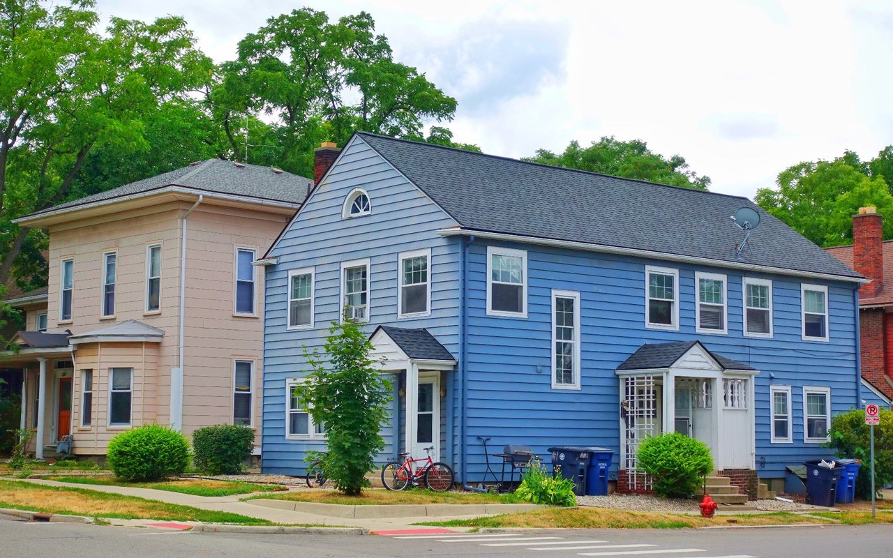 Blue two-story house with white trim and a small covered entry on a tree-lined street.