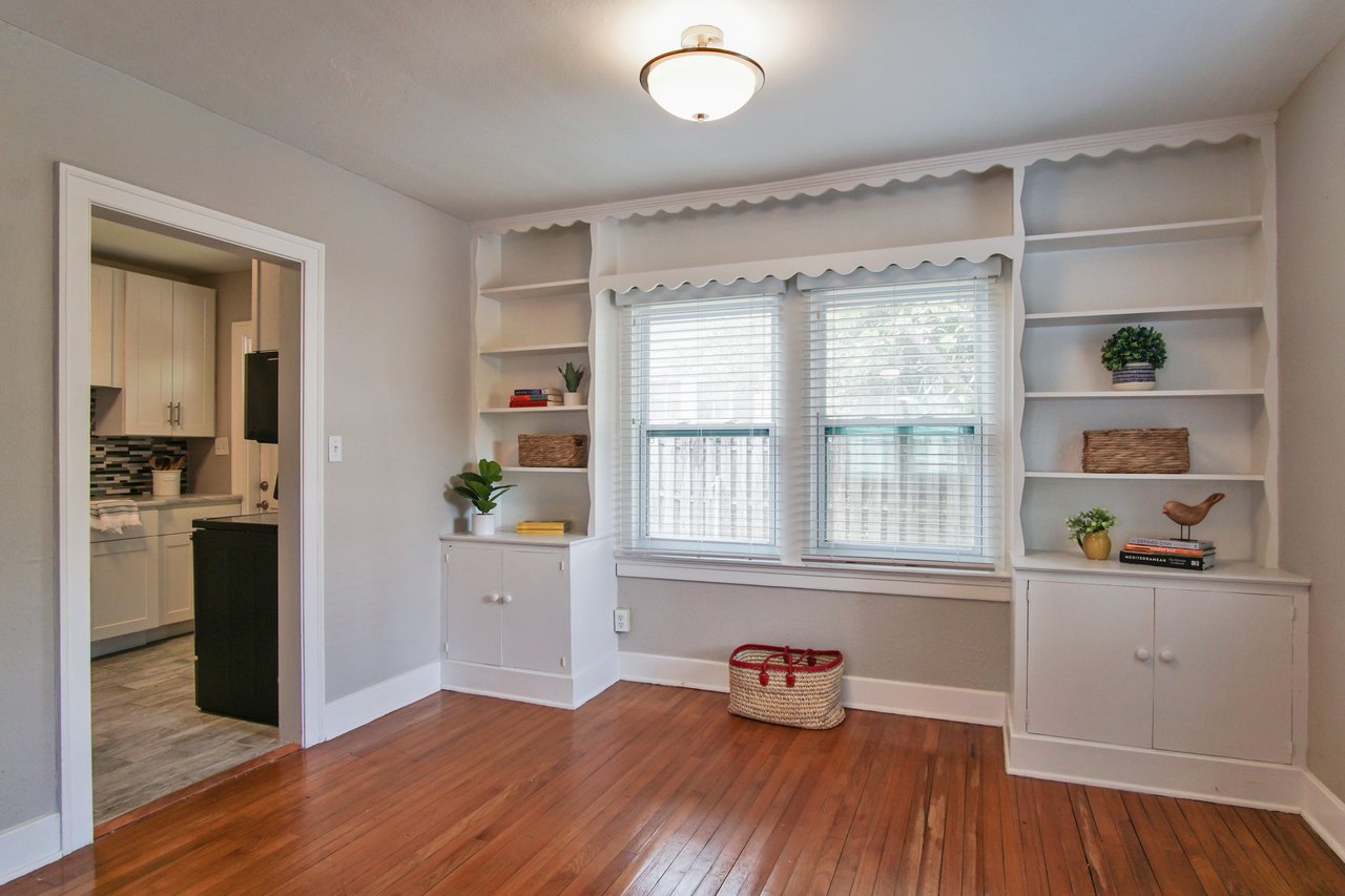 A bright room with wooden floors and built-in white shelves surrounding a window. Shelves hold plants, baskets, and books. Adjacent is a modern kitchen.