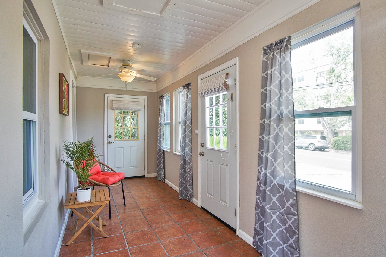 Bright sunroom with terracotta tiles and beige walls. A ceiling fan hangs above. A potted plant on a wooden table and a red cushion on a chair add warmth.