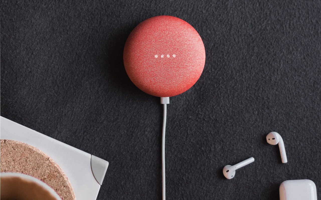 Coral-red fabric smart speaker with four white indicator lights, beside wireless earbuds.