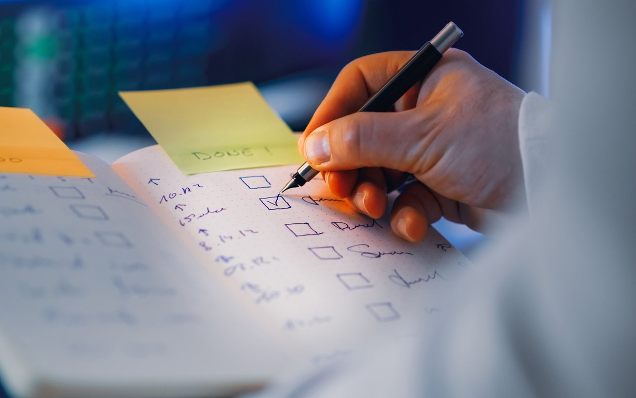 A hand checks off items on a handwritten to-do list beside a yellow sticky note.