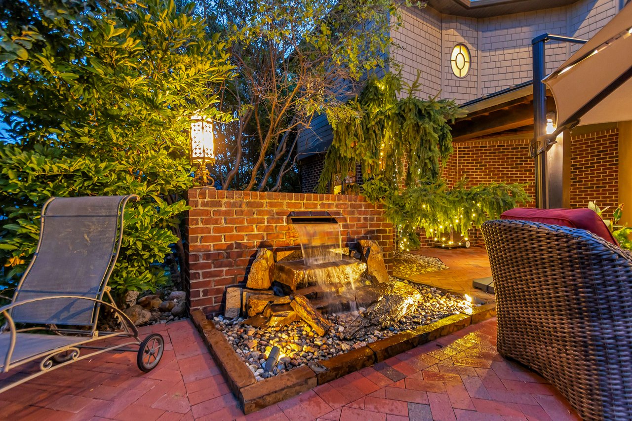 Brick patio with a tiered fountain, outdoor seating, and warm lights among dense greenery.