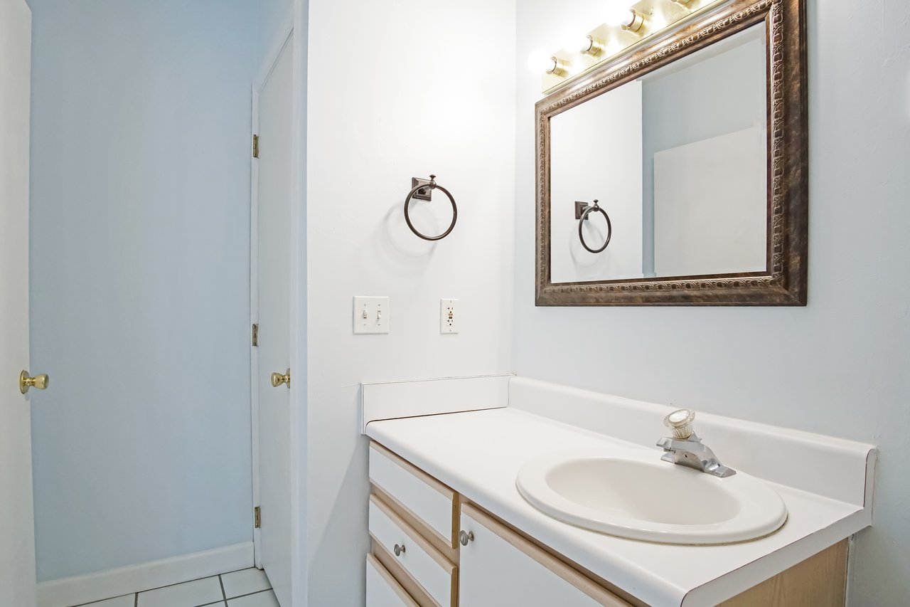 Clean bathroom with white walls, wooden-framed mirror, and bright lights. Simple vanity with beige counter and sink, creating a fresh, minimalist tone.