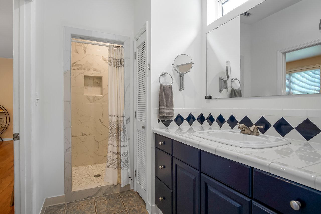 Bright bathroom with navy and white vanity on the right, large mirror above. Doorway leads to shower with beige walls. Modern and clean design.