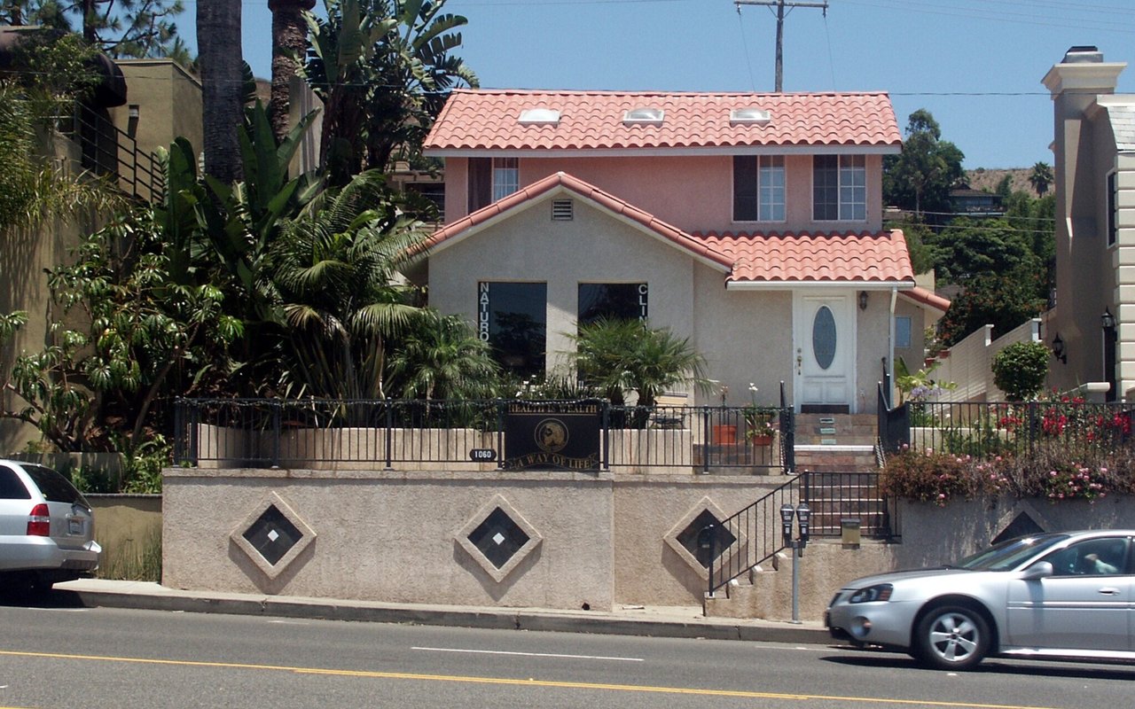 Two-story stucco building with red tile roofs, tropical landscaping, and business signage behind a decorative wall.