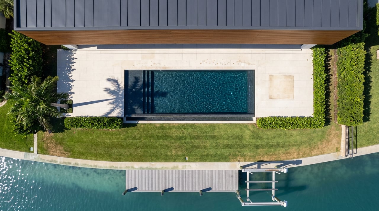 Overhead waterfront estate with a rectangular pool, striped lawn, concrete seawall, and teak dock extending into blue-green water.