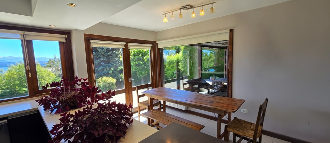 Dining area with a long wooden table and broad windows overlooking trees, a lake, and distant mountains.