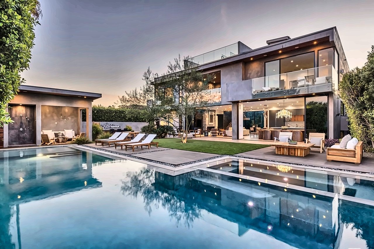 Modern glass-fronted home beside a reflecting pool, with landscaped lawn and outdoor seating at dusk.