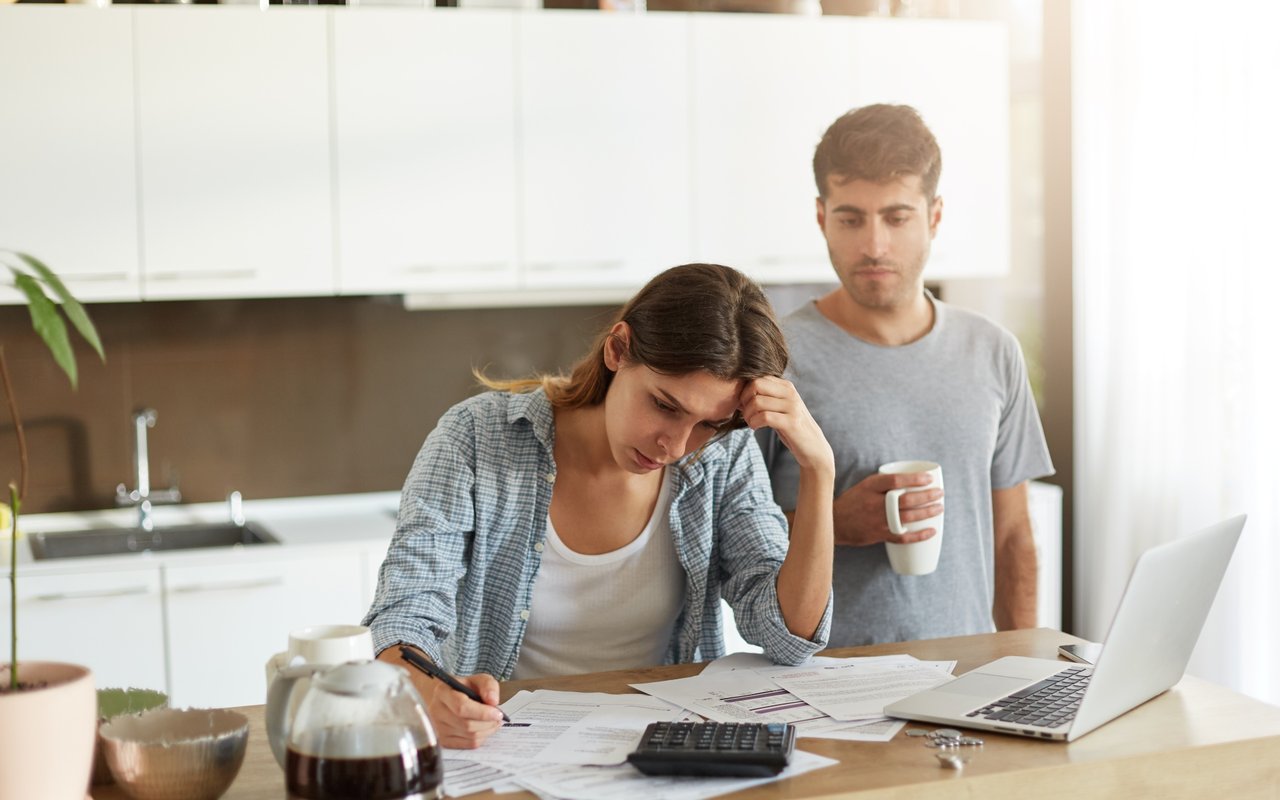 A worried woman reviews paperwork at a kitchen table as a man stands behind her with coffee.