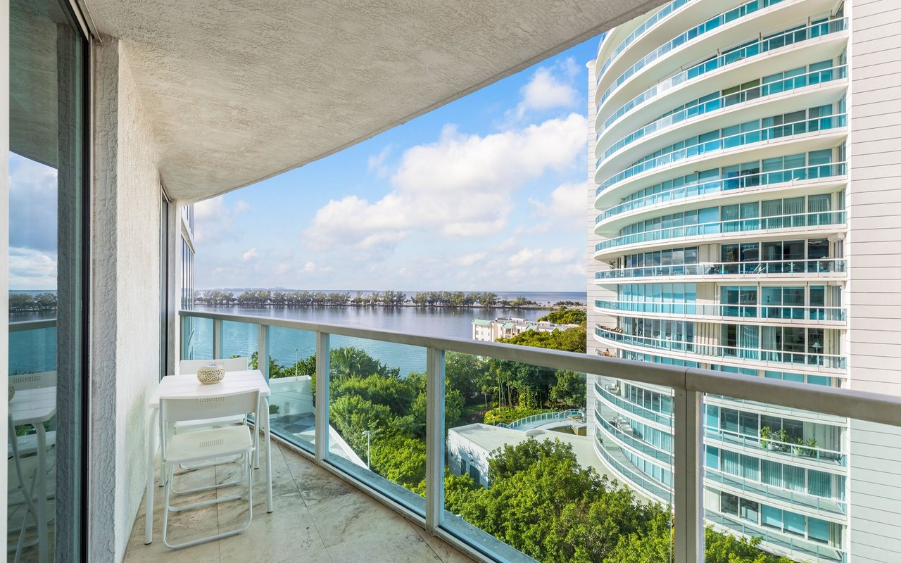 Condo balcony with white patio table and chairs overlooking the water and a neighboring high-rise.