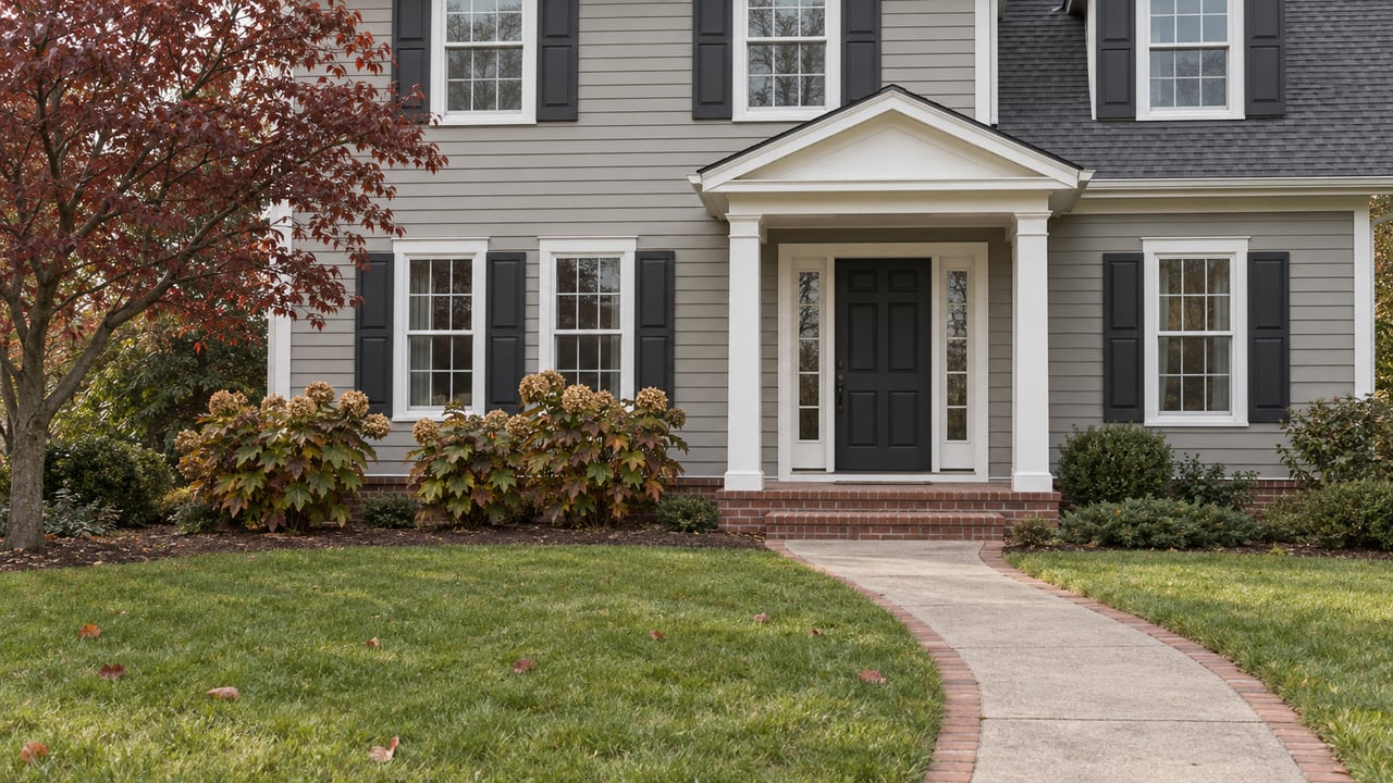 Traditional gray lap-sided home with white entry columns, brick steps, a deep front lawn and burgundy dogwood foliage.