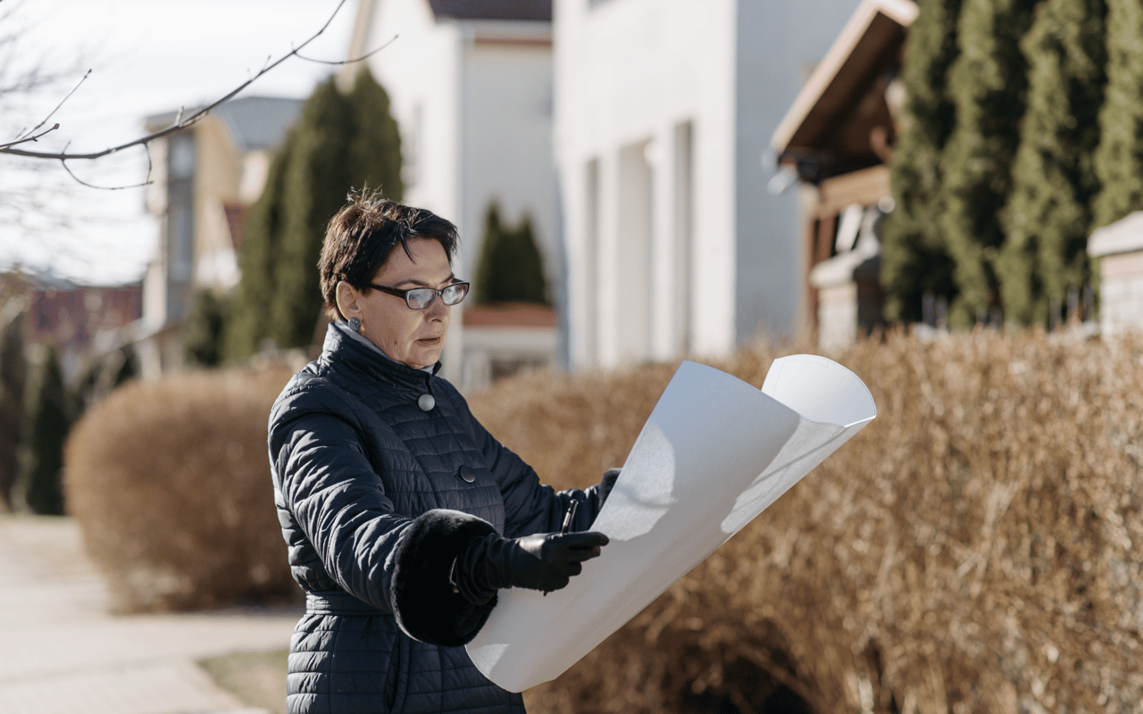 Woman in glasses reviews large building plans beside hedges and homes.