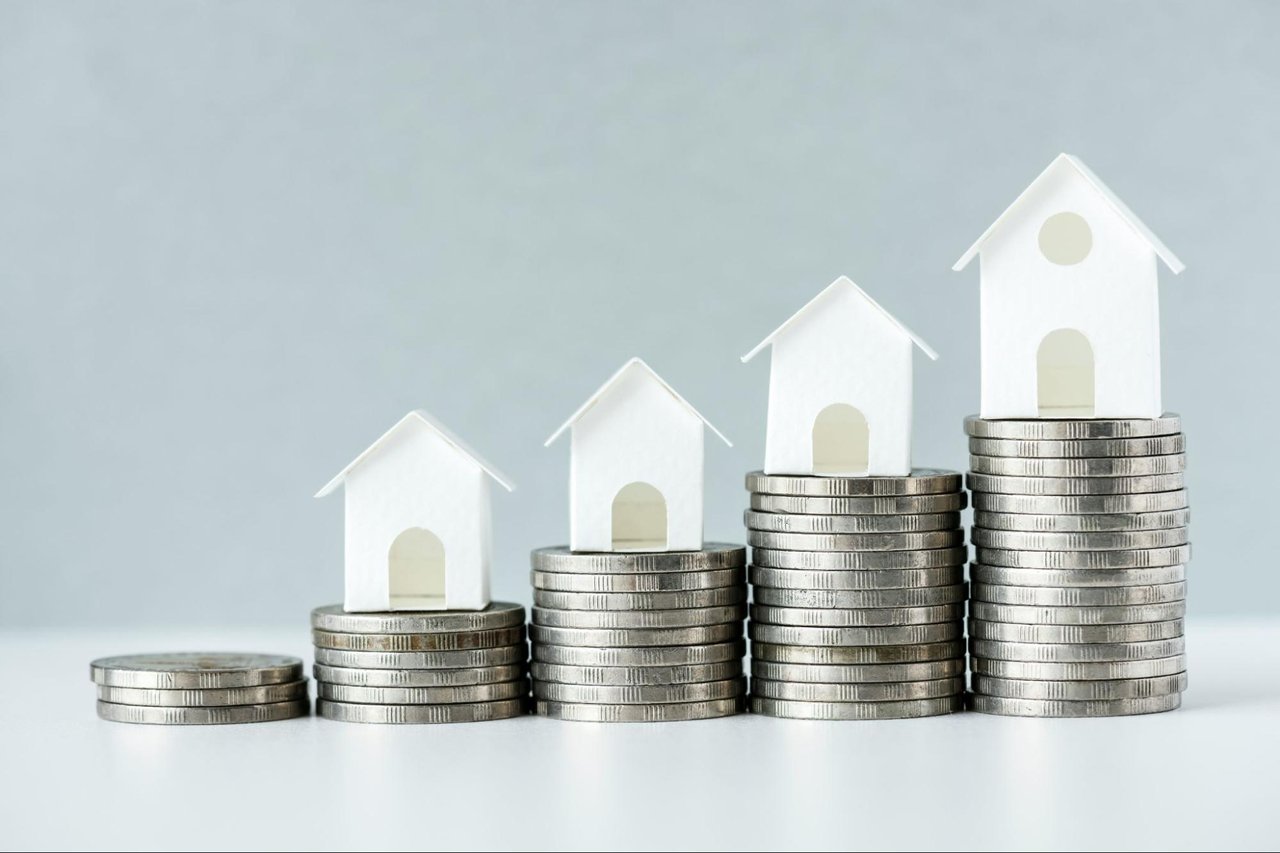 White model houses stand on increasingly tall stacks of coins, suggesting rising real estate prices.