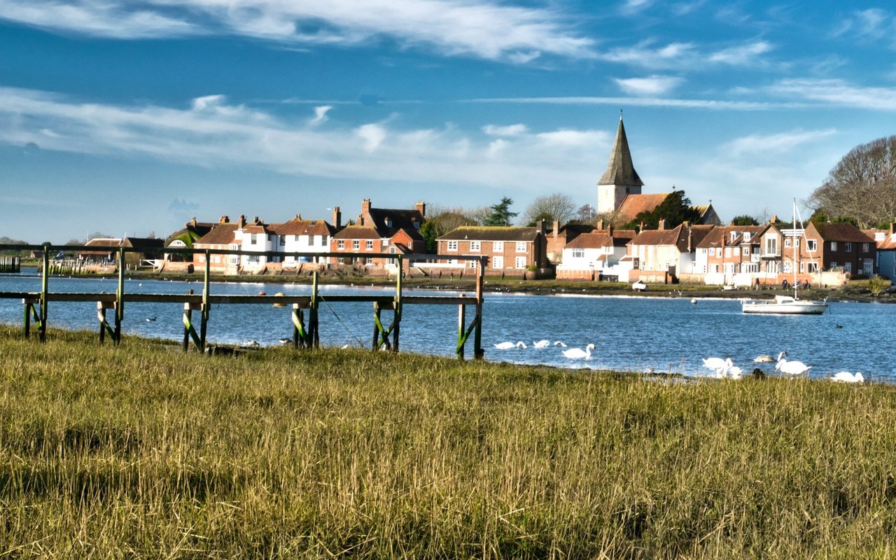 Waterfront village with a church steeple, grassy shoreline, swans, and boats.