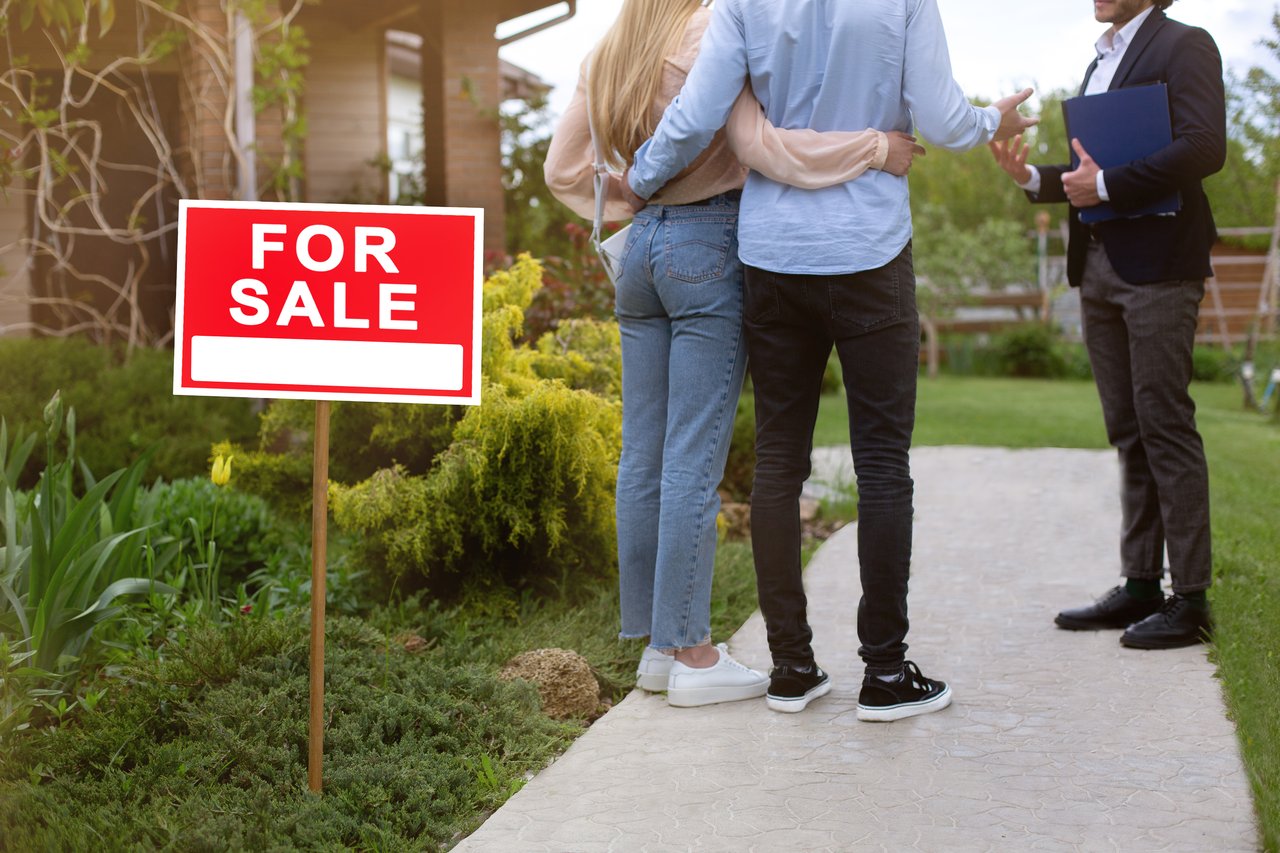 Couple speaking with a real estate agent outside a home with a red For Sale sign.