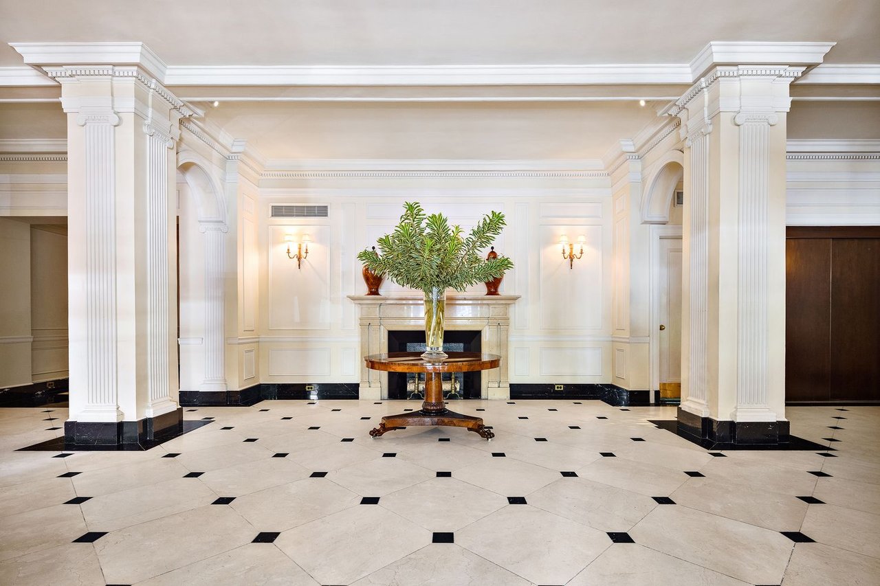 Residential lobby with ornate white columns, a fireplace, and a central table with a leafy arrangement.