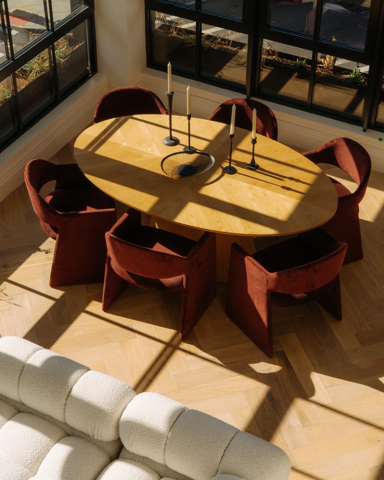Sunlit dining area with an oval wood table, rust-red chairs, and tall black-framed windows.