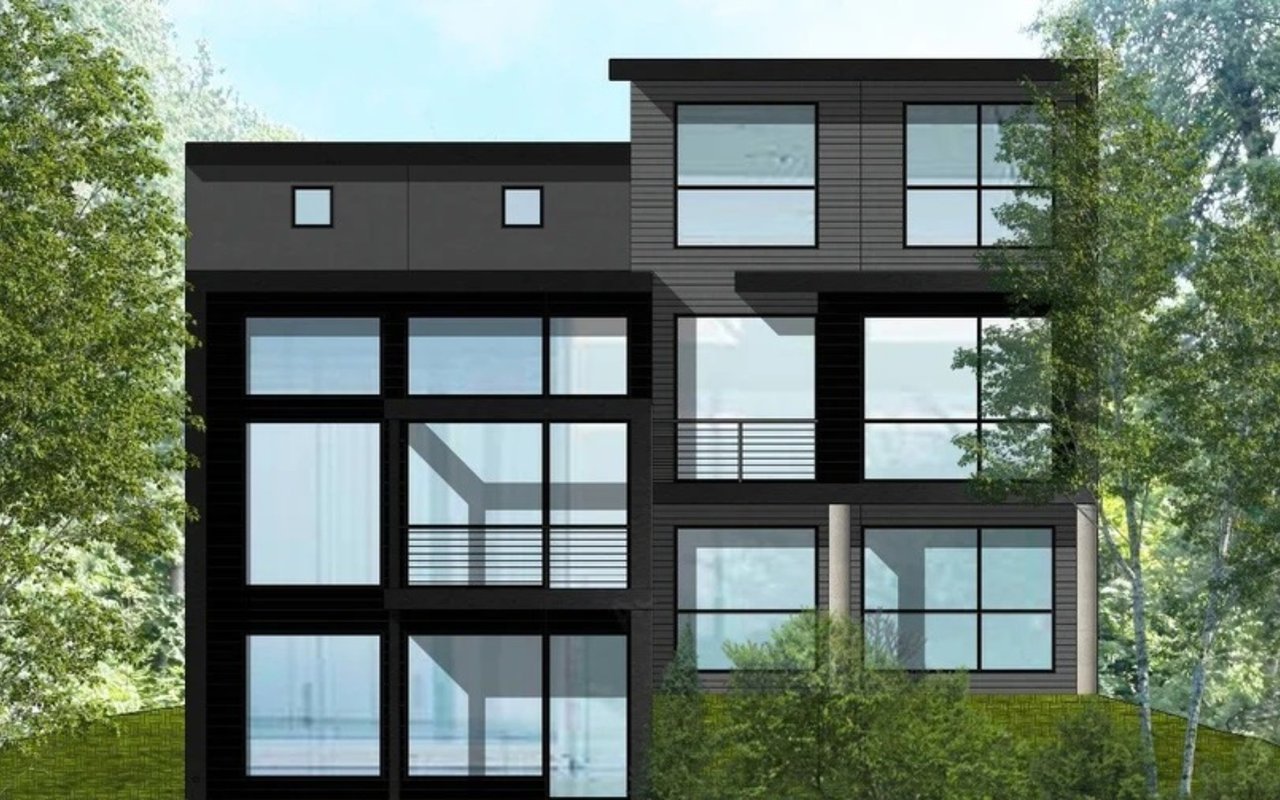 Modern lakefront home with dark siding, expansive grid windows, and a balcony among trees.