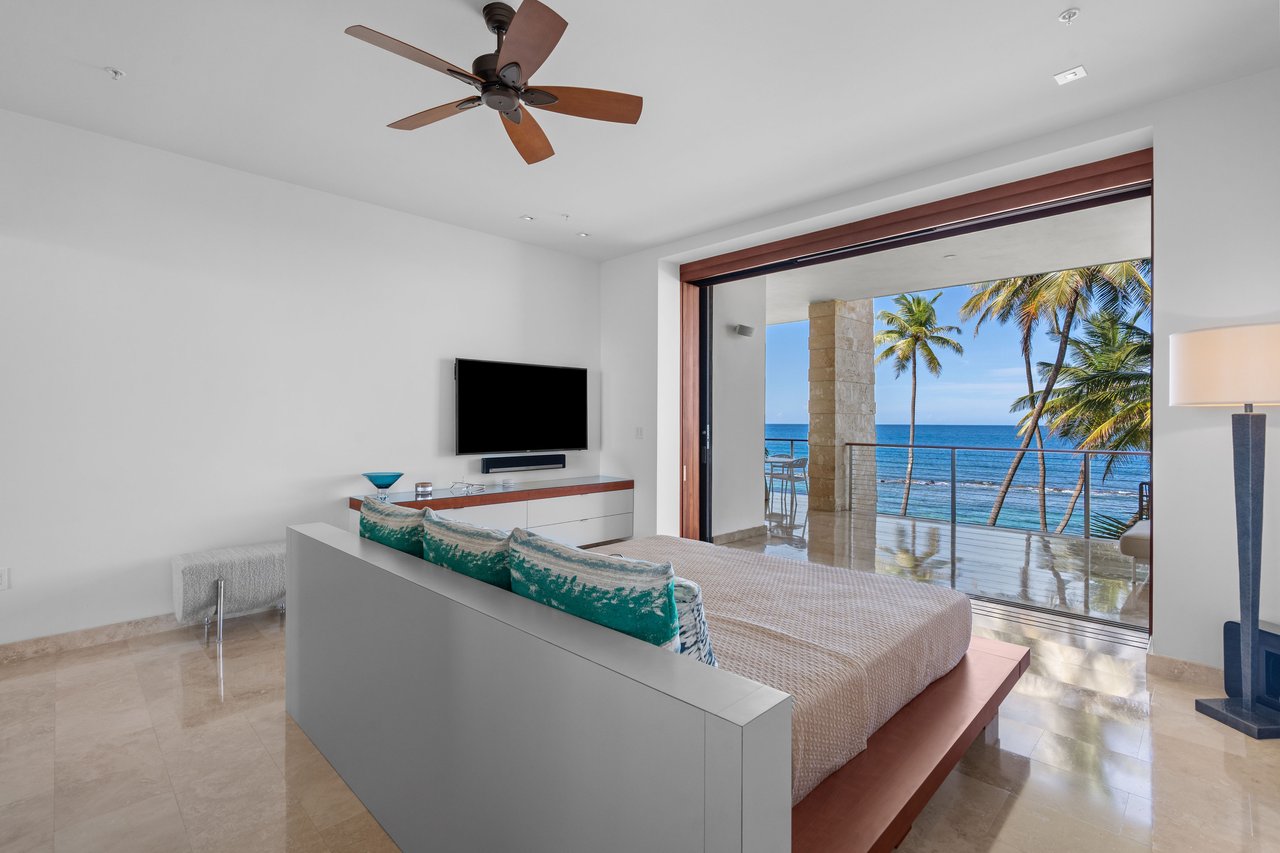 Bedroom with turquoise pillows and open balcony doors overlooking the ocean and palm trees.