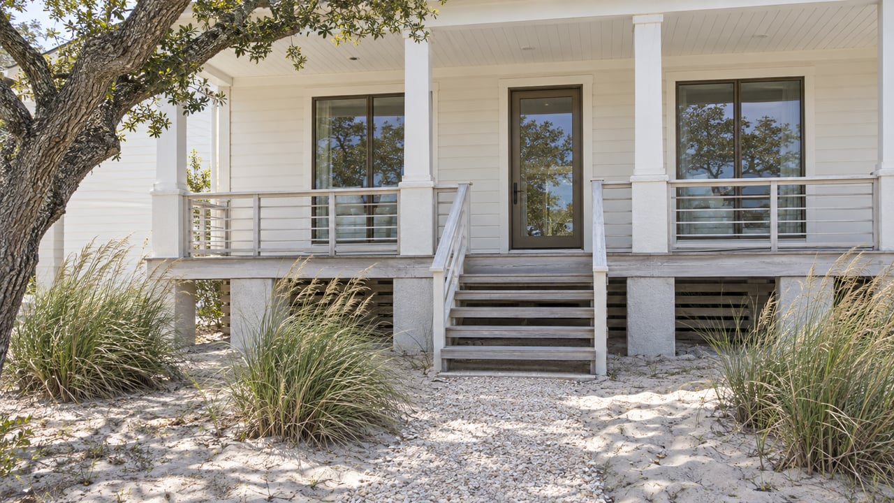 Raised coastal house with ivory siding, timber entry steps, and dune grass in a sandy front yard.