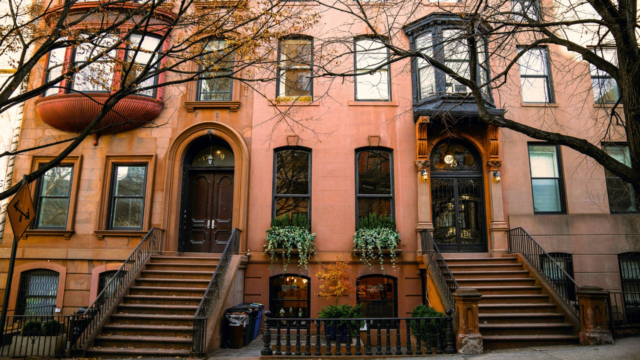 Adjoining pink-brownstone townhouses with tall stoops and ornate entrances beneath bare tree branches.
