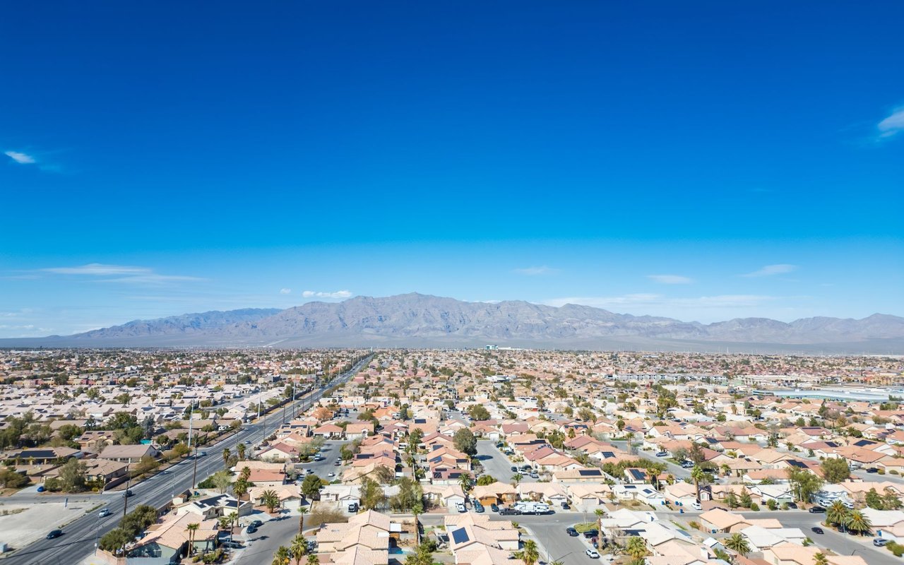 Aerial view of a desert suburb with low-rise homes, a broad road, and mountains beyond.
