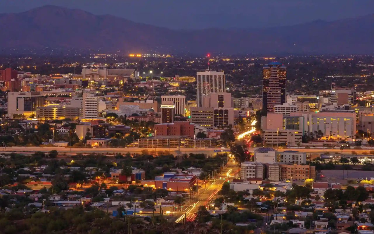 Aerial dusk view of downtown Tucson looking east toward the Rincon Mountains with glowing streetlights and traffic headlights on Broadway Blvd.