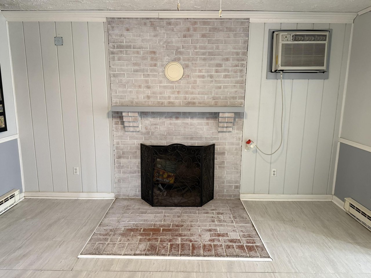 Brick fireplace with tiled hearth and black screen in a vacant room with a window air conditioner.