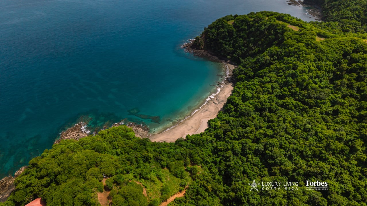 Aerial view of a forested coastal lot beside a turquoise cove and sandy beach.