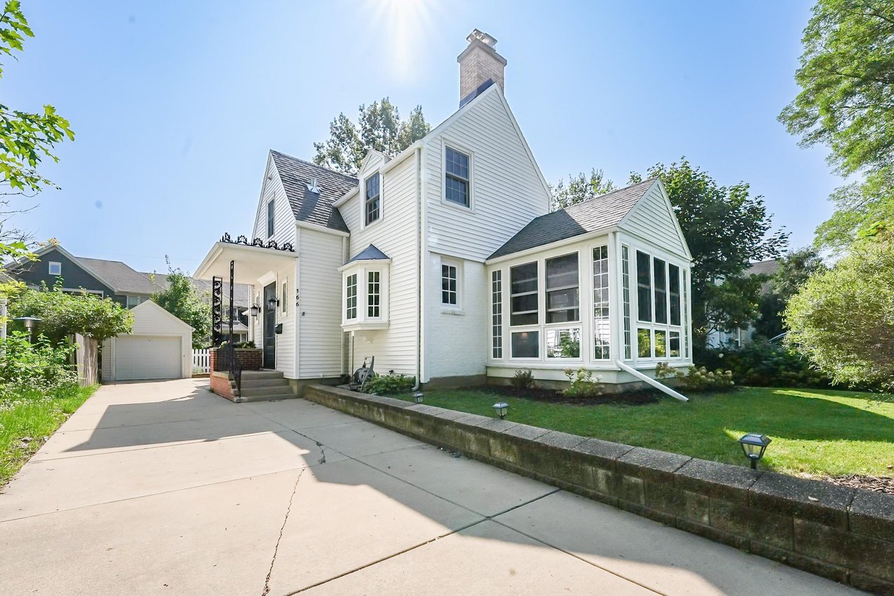 A white older two-story home with a brick chimney, dormers, and an enclosed sunroom on a sunny day in an established Madison neighborhood.