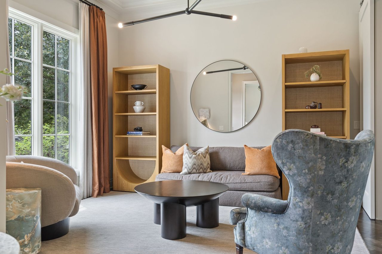 Living room with a round wall mirror, built-in wood shelving, and a floral wingback chair, furnished by Ashby Collective, in the Tarrytown home at 2600 Westover Road, Austin.