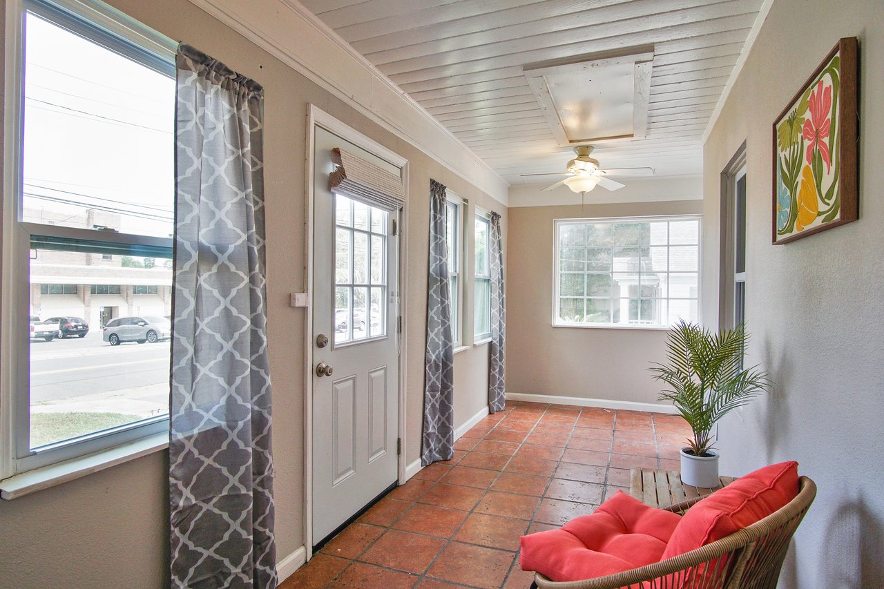 Bright sunroom with terracotta tiles, large windows with gray patterned curtains, a white ceiling fan, red cushioned chair, and a potted plant.