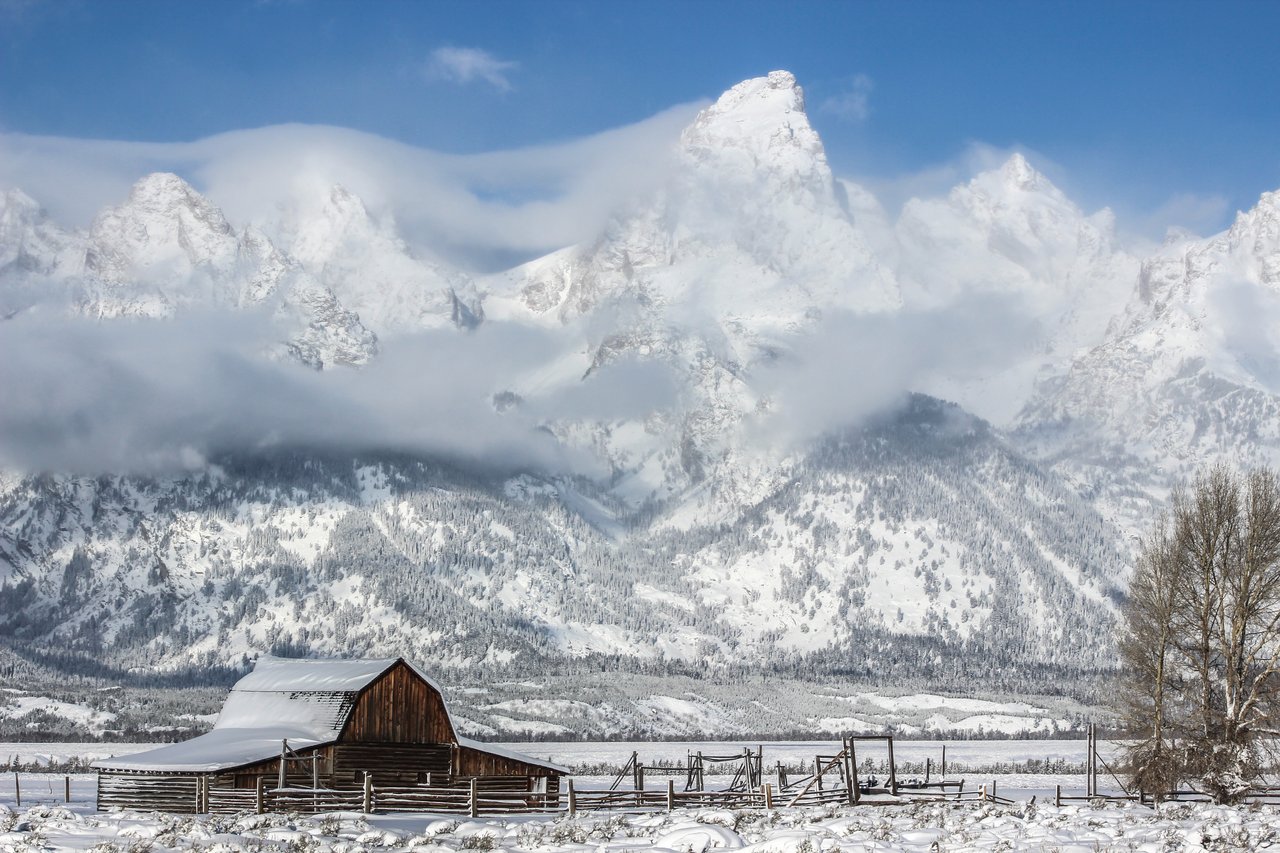 A winter photograph of the Mormon Barn in Grand Teton National Park, with a backdrop of the Tetons covered in snow with some cloud cover.